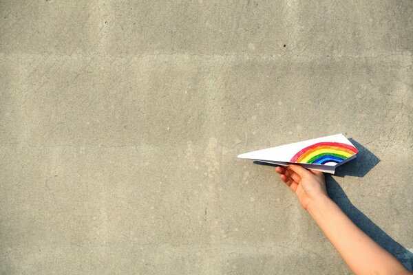 Avión de papel con ala de arco iris contra fondo de pared blanca