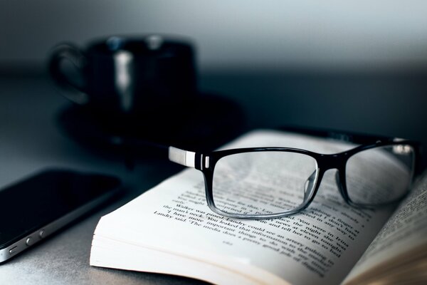 Una taza negra junto a un libro, gafas y un iphone 4