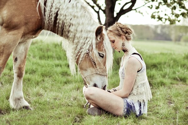 The girl is sitting on the grass and feeding the horse