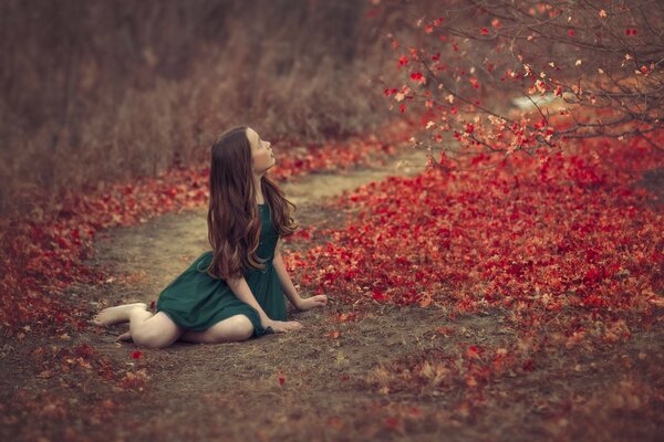 The girl sits on the road and looks at the red fallen leaves