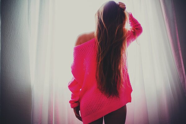 A girl in a pink sweater against the background of curtains and light