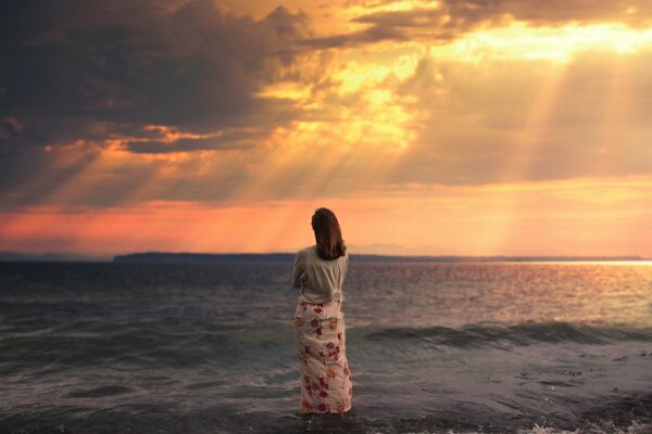 A girl in a long skirt looks at the sunset at the sea