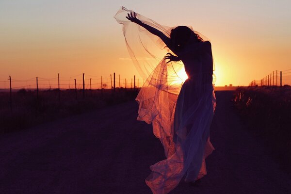 Ragazza snella in abito di garza che balla sulla strada sullo sfondo del tramonto serale