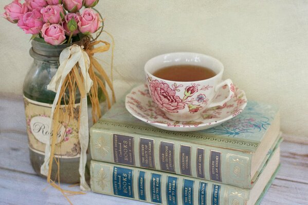 A bouquet of pink roses in a jar and books lying next to a cup of tea