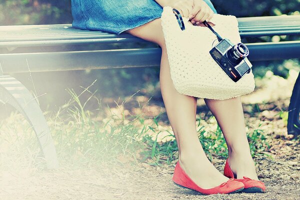 Beine in roten Ballerinas eines Mädchens auf einer Bank in einem Jeansrock mit einer Kamera und einer Stricktasche in den Händen