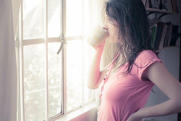 A brunette girl with wet hair in a pink T-shirt drinks from a mug by the window