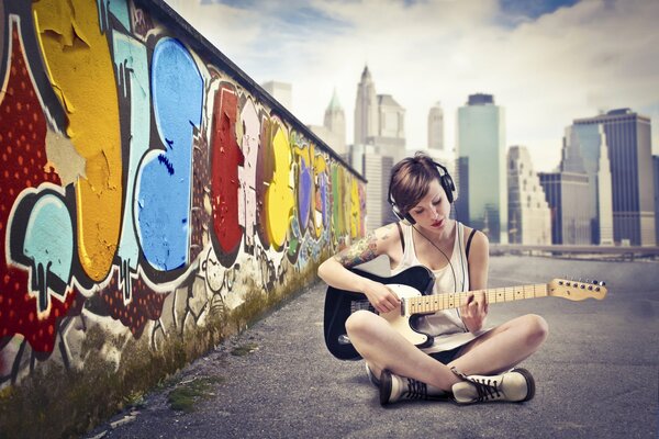 A girl with headphones against a graffiti wall plays guitar