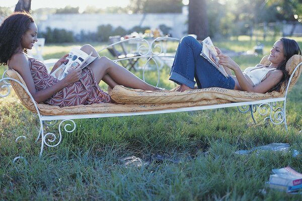 Two girls are lying on a bench and reading magazines