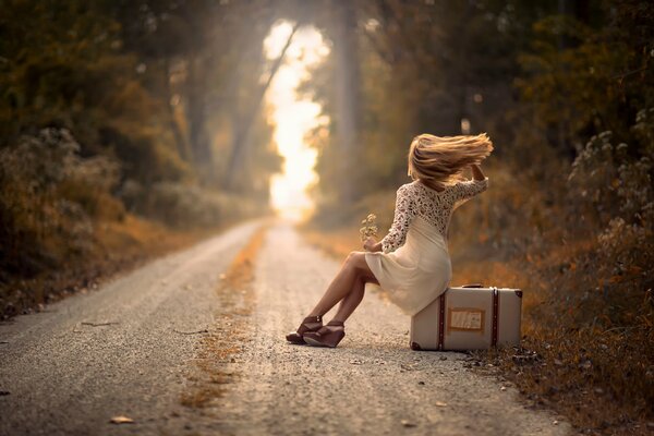 A girl sitting on a suitcase on the side of the road. The wind blows her hair