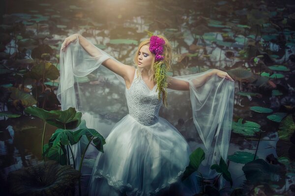 Una chica con un vestido blanco en un estanque con nenúfares con una flor brillante en el pelo