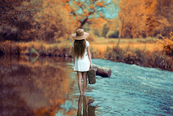 Una chica con un vestido corto blanco y un sombrero camina por el río con una maleta, vista trasera