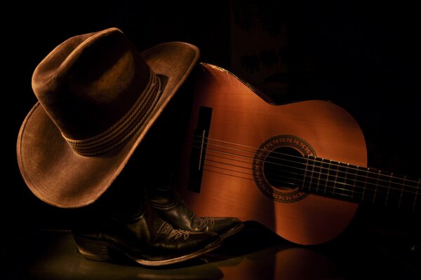 Cowboyschuhe und Hut liegen neben einer Gitarre auf dunklem Hintergrund