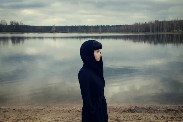 Foto drammatica di una ragazza in un cappotto nero su uno sfondo di un lago e una foresta grigia senza fogliame