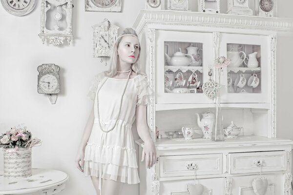 A girl is a porcelain doll on a background of white objects and a clock