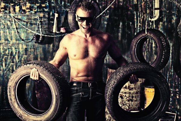 A man with black glasses and a bare torso holds tires in his hands