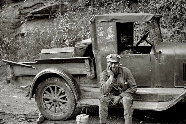 An old truck, and a man is sitting on the doorstep with his head propped up