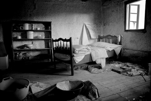 A house abandoned by people with broken windows, an unmade bed and shelves with the remaining dishes in black and white