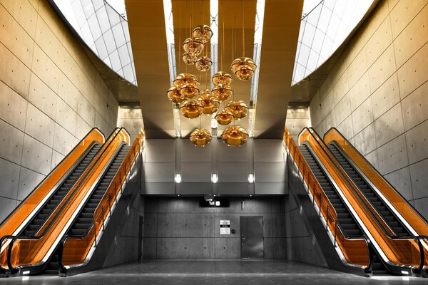 Escaliers mécaniques orange et lustre orange en forme de fleurs dans une pièce bordée de gros Carreaux gris