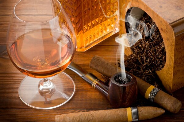 On the table there is a glass of cognac and cigars and a pipe