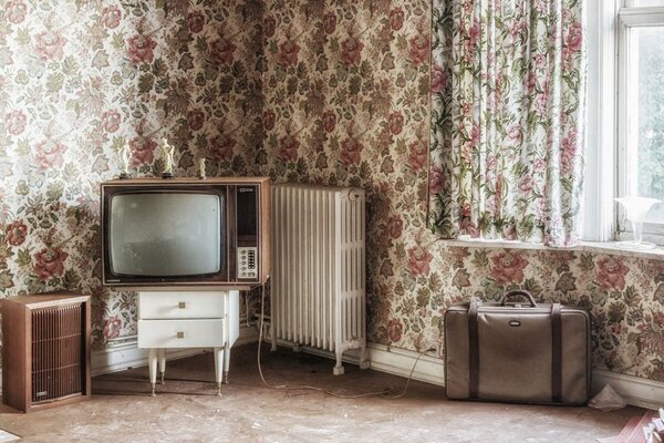 A room with floral wallpaper. TV and suitcase