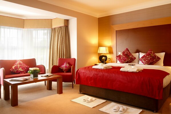 A modern-style bedroom with a double bed under a red bedspread and a seating area in the bay window