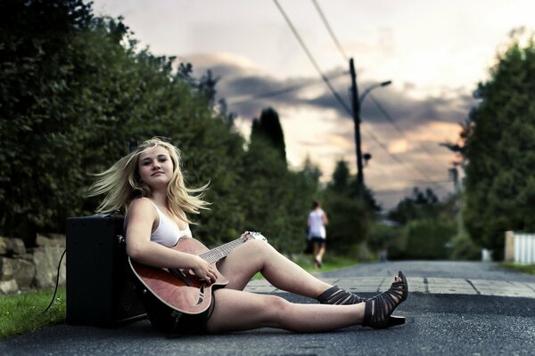 A girl with a guitar is sitting on the asphalt