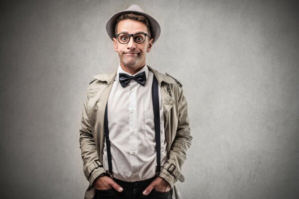 A man in suspenders, a hat, glasses and a coat is standing near a gray wall