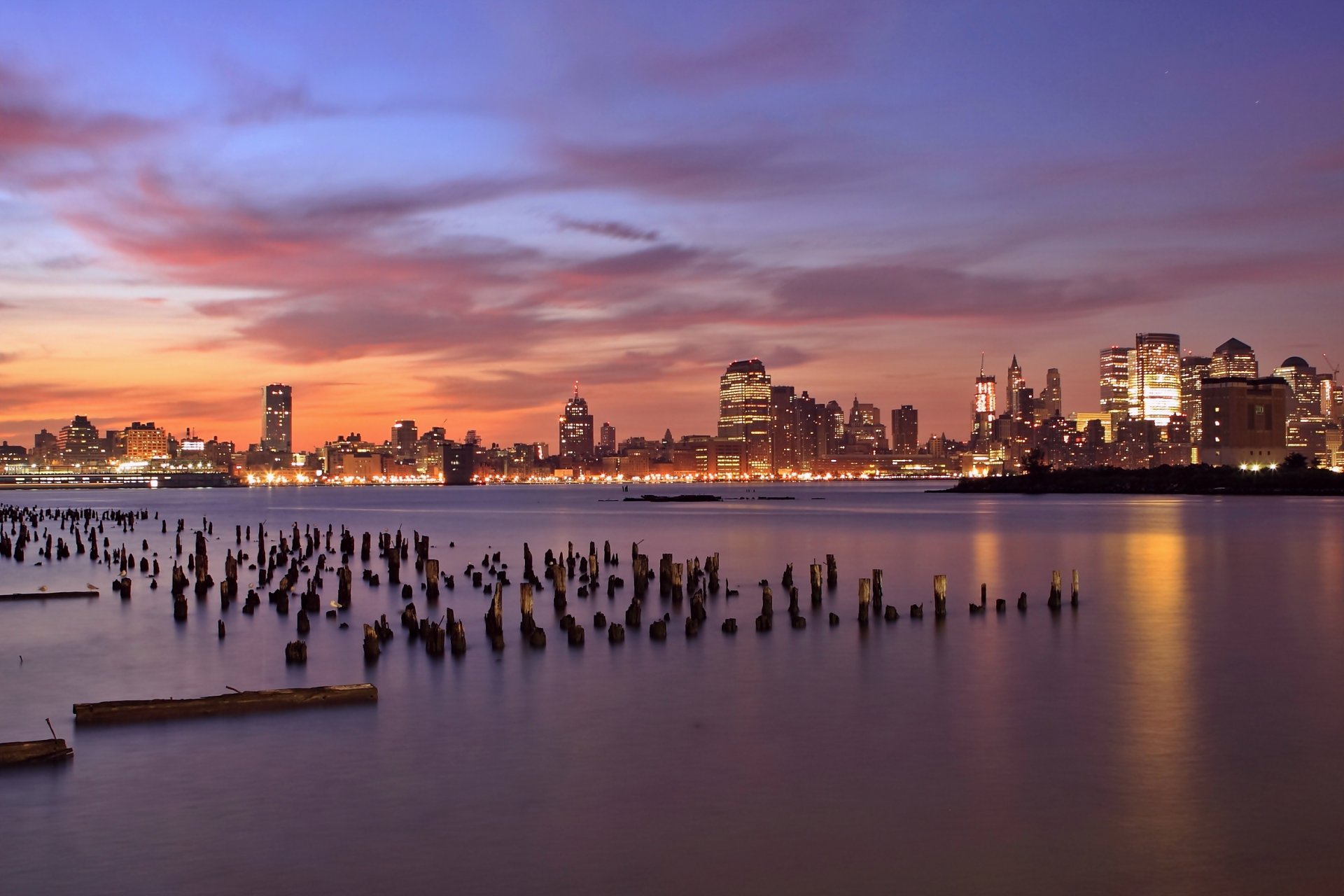 estados unidos jersey city nueva jersey nueva jersey noche naranja puesta de sol lila cielo nubes rascacielos iluminación luces río hudson madera bolardos