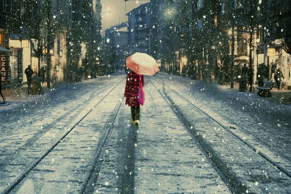 Das Mädchen im roten Mantel geht die Straßenbahngleise entlang. sich unter einem Regenschirm vor nassem Schnee verstecken