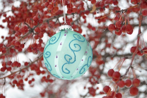 Boule de Noël turquoise avec des motifs suspendus sur des fils blancs sur fond de branches avec de petites pommes rouges en hiver