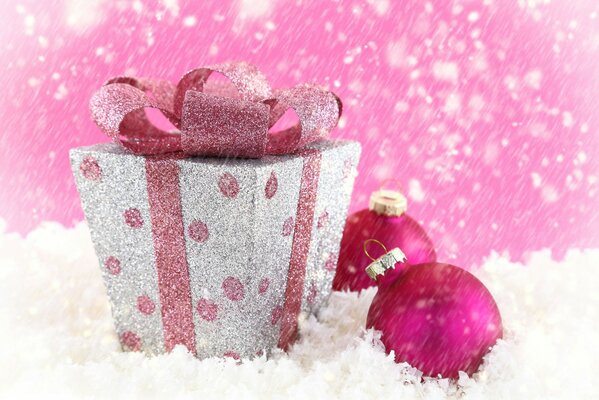 In the snow, a box with a pink ribbon, next to two pink Christmas balls and falling snow