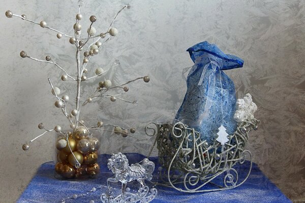 Un caballito de cristal, una bolsa azul atada con una cinta en un trineo, cuentas blancas, instalación de un árbol de Navidad en un frasco con bolas doradas de año nuevo sobre un mantel azul y blanco contra una pared gris