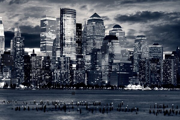 Black-and-white, panoramic photograph of a megalopolis, high-rise buildings illuminated by lights on the evening sky against the background of the ocean