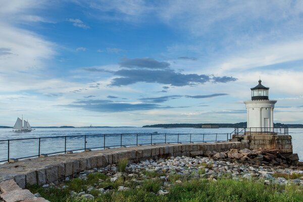 Calm in the sea, a white lighthouse on the shore, and in the distance a ship with white sails