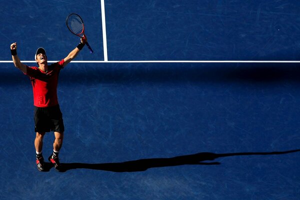 A tennis player in a red jersey rejoices at the victory on the court, raising his racket up