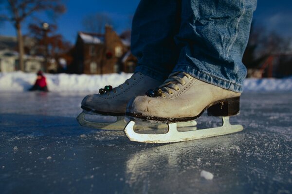 Mann in Schlittschuhen und Jeans auf einer Eisbahn in der Nähe des Hauses. Im Hintergrund sitzt ein Mann in einer roten Jacke auf dem Eis