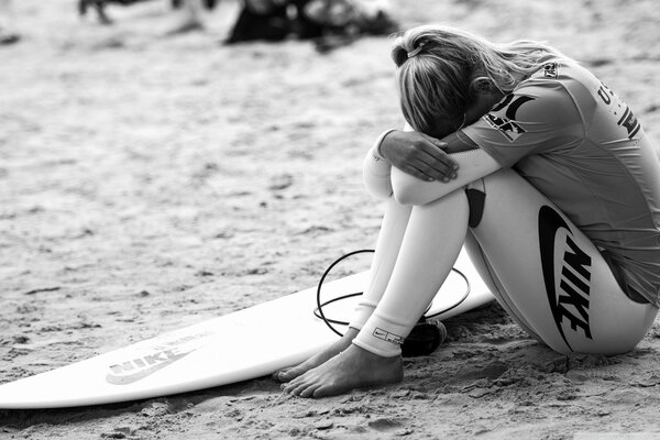 Black and white photo of a girl with her head buried in her knees with a surfboard lying on the sand