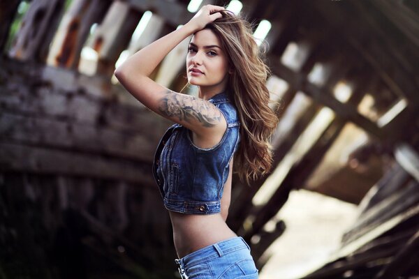 A girl with long hair in jeans and a denim vest with a tattoo on her arm