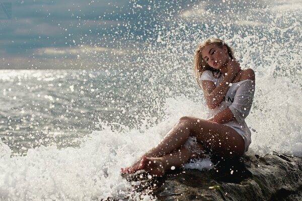 A young blonde in white clothes is sitting on the seashore and splashing