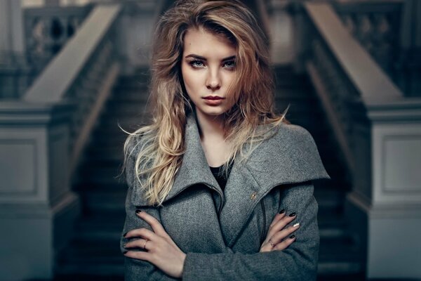 A girl in a pose with her hands on her sides looks forward against the background of a white stone staircase