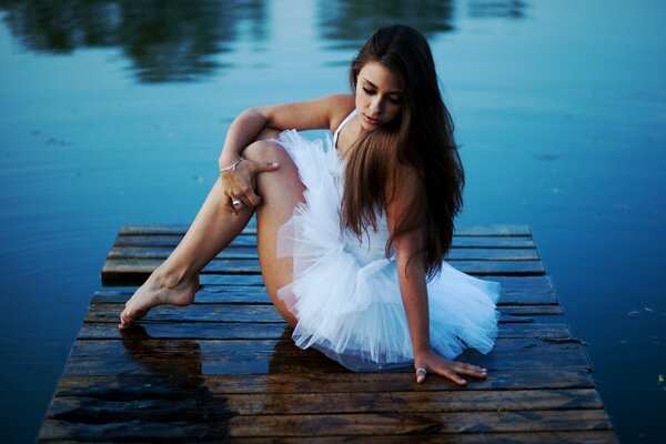 The ballerina girl is sitting by the lake on the pier