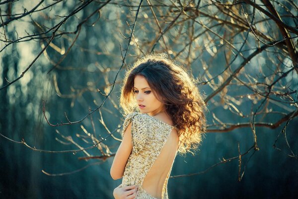 A girl in a dress with a large neckline on the back on a blurry background of branches without leaves