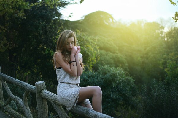 A blonde girl in a white dress is sitting on a fence against a background of trees