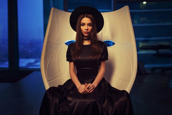 A girl in a black dress and hat is sitting on a chair