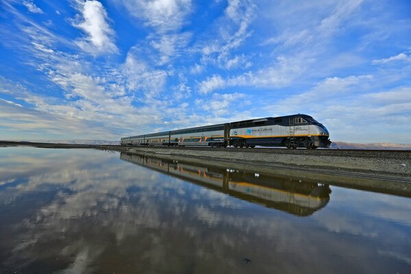 Un tren viaja a lo largo de un lago en el que se reflejan las nubes