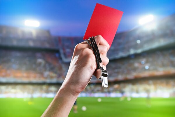 The hand of a football referee with a red card and a whistle