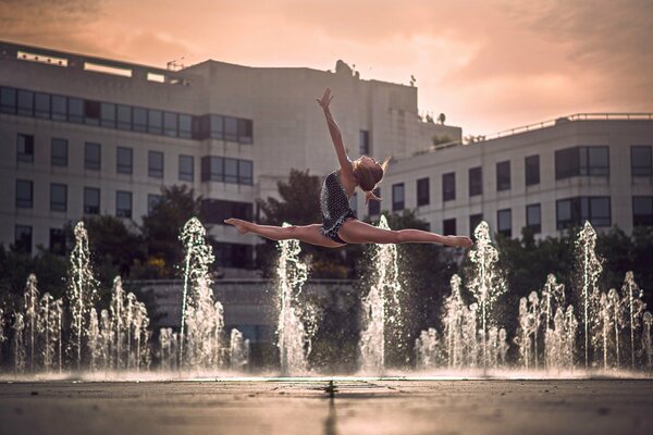 A girl in flight does the splits against the background of fountains and buildings