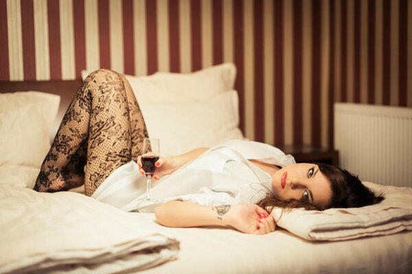 A brunette girl is lying on a white sheet, holding a glass of red wine in her hands