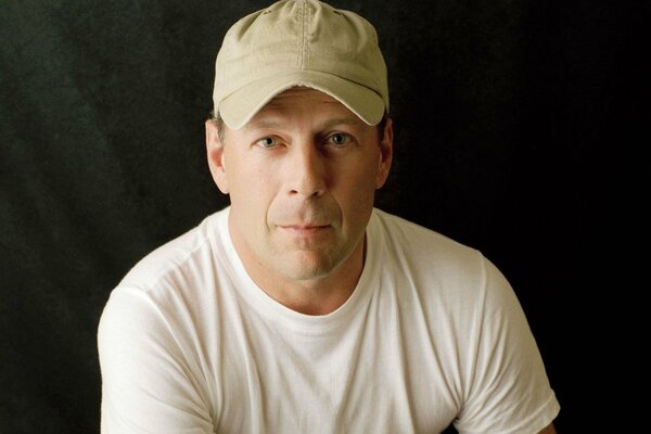 Bruce Willis is sitting in a beige T-shirt and a cap