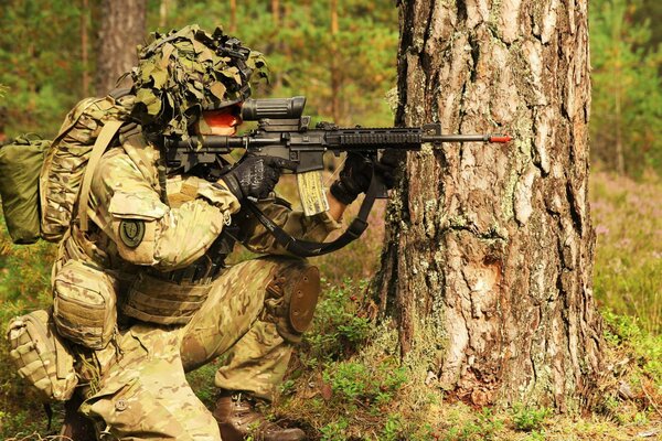 A soldier is sitting behind a large tree with a gun cocked